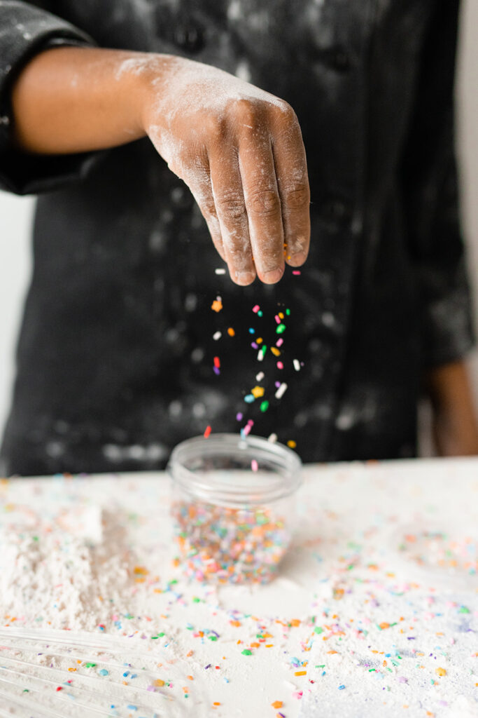 A black custom cake maker covered in flour, sprinkling sprinkles in Guelph Ontario