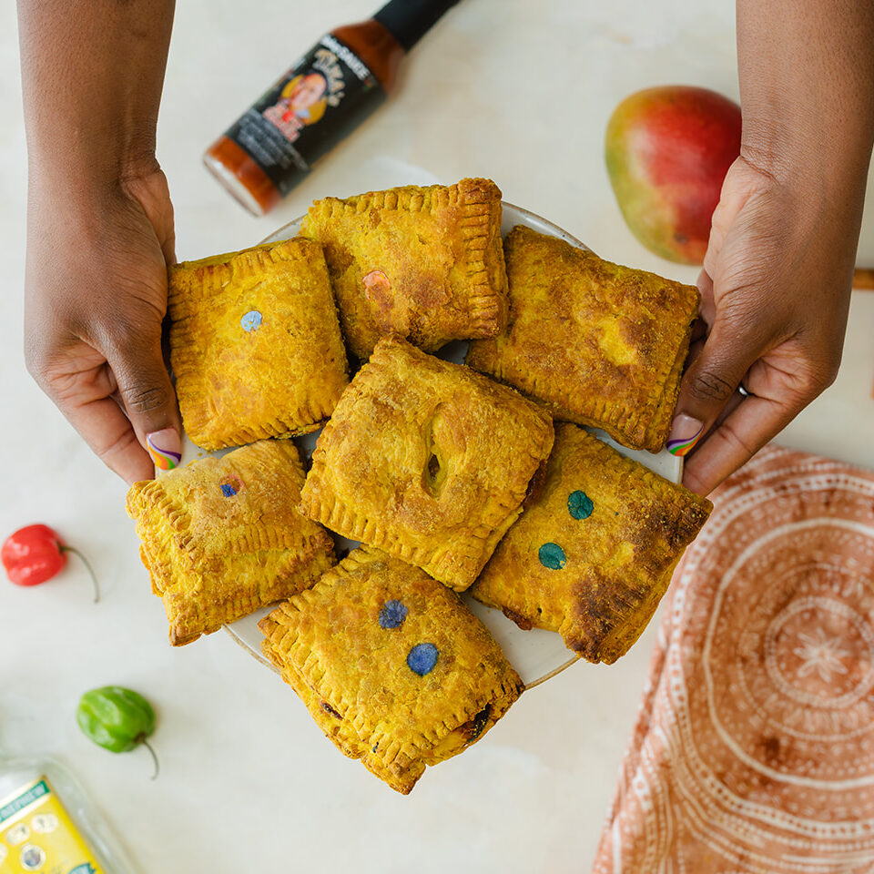 A pair of hands holding a platter of Jamaican patties above a counter with scotch bonnet peppers, a rum bottle, mangoes, hot sauce and a decorative napkin.