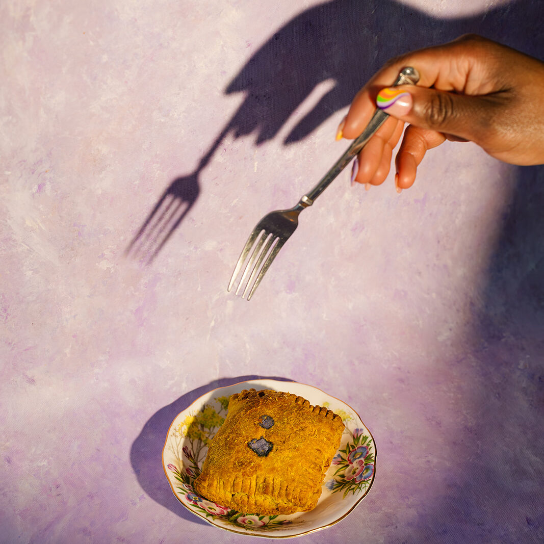 A hand holding a fork above a Jamaican patty on a small decorative plate with a purple background in Kitchener ON